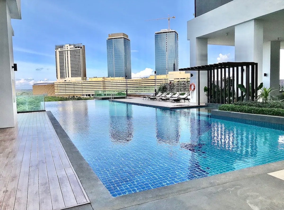 An expansive swimming pool reflects the clear blue sky, framed by modern high-rise buildings. Sun loungers are arranged around the pool area, complemented by green landscaping. The overall ambiance suggests a serene and inviting space for relaxation.