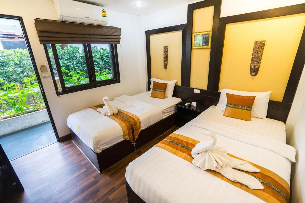 A clean and well-lit guest room features two spacious beds, each adorned with decorative pillows. Soft, neutral tones are complemented by warm wooden flooring. Natural light enters through a large window, which overlooks a green outdoor space, creating a calming ambiance.