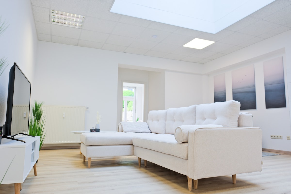 A spacious living area features a light-colored sectional sofa and a flat-screen TV. Natural light enters through a skylight, illuminating the neutral color palette. Two framed prints on the wall display calming landscapes, contributing to a serene environment.