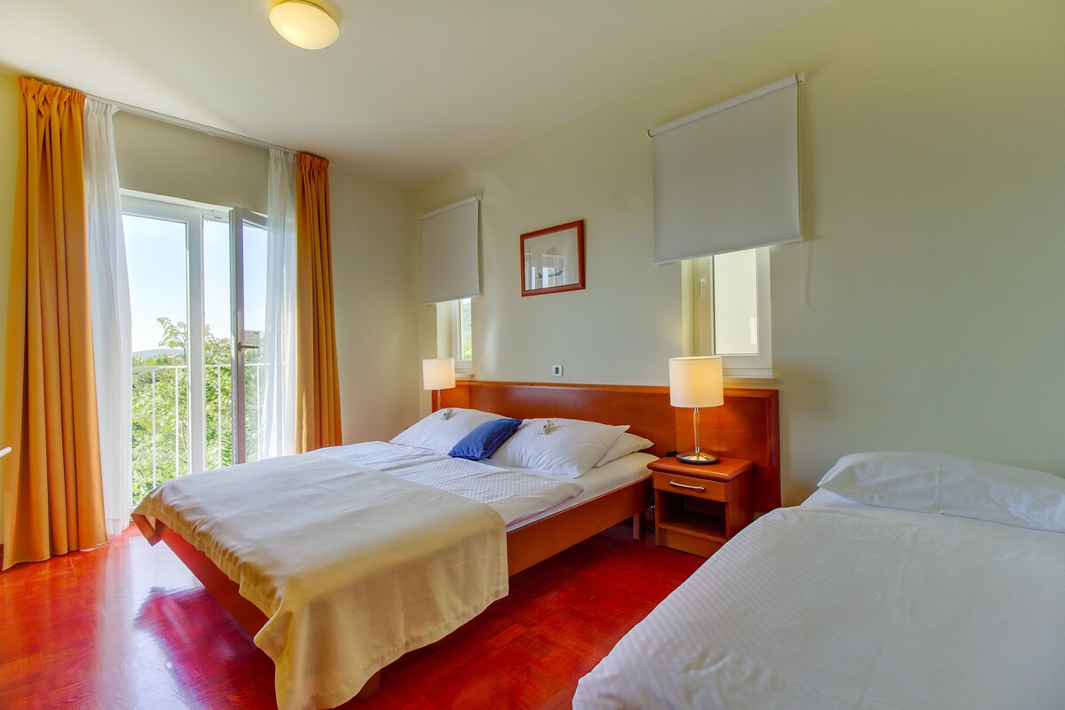 A bright sleeping area features two spacious beds with white linens. Natural light filters in through two large windows adorned with light drapes. A bedside table with a lamp complements the wooden headboards, creating a serene and relaxing environment.