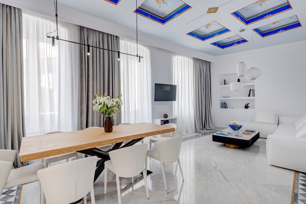 A spacious dining and living area features a wooden table surrounded by white chairs. Large windows with sheer curtains allow ample natural light. A modern sofa is positioned opposite a wall-mounted television, with decorative shelving displaying books and a vibrant centerpiece on the table.