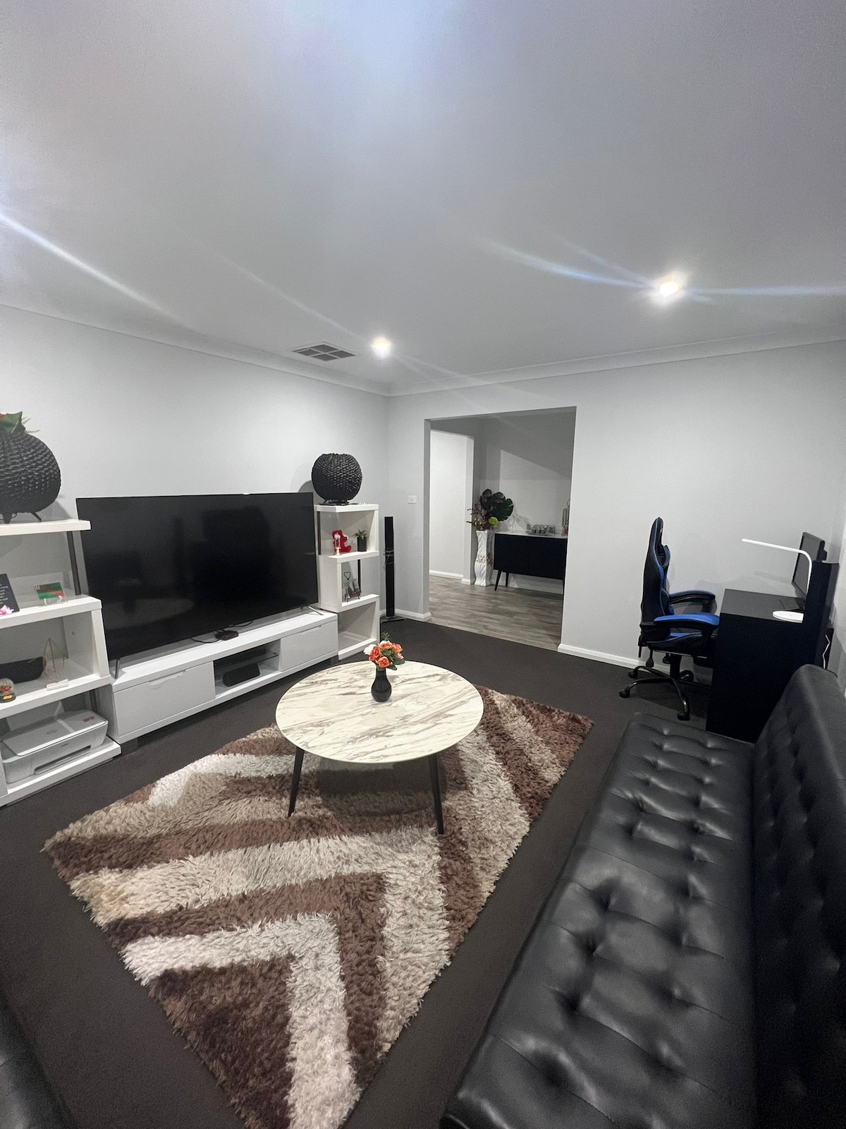 A modern living area features a large, flat-screen TV mounted on a white entertainment unit. A round, marble coffee table sits on a geometric-patterned rug, surrounded by dark sofas. A doorway leads to a secondary space, which can be seen in the background.