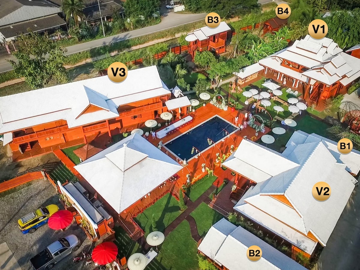 An aerial view of the property showcases multiple structures with traditional Lanna style architecture and bright red accents. A central pool area is surrounded by lounge chairs and umbrellas, while pathways lead to the villas and bungalows nestled among well-maintained greenery.
