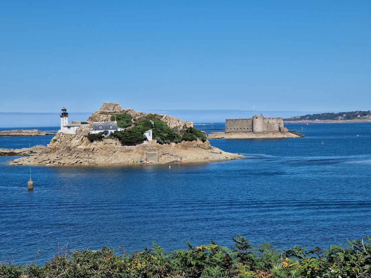 A scenic view of an island featuring a lighthouse and a historical fort, surrounded by calm blue waters. The sunlight highlights the natural landscape, and a few small boats are visible in the distance, contributing to the serene atmosphere of the coastal scene.