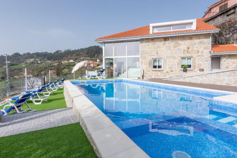 A modern stone house with large glass windows is seen beside a rectangular swimming pool. Lounge chairs are arranged around the pool area, overlooking a green landscape. A white umbrella provides shade to the seating area, enhancing outdoor relaxation.