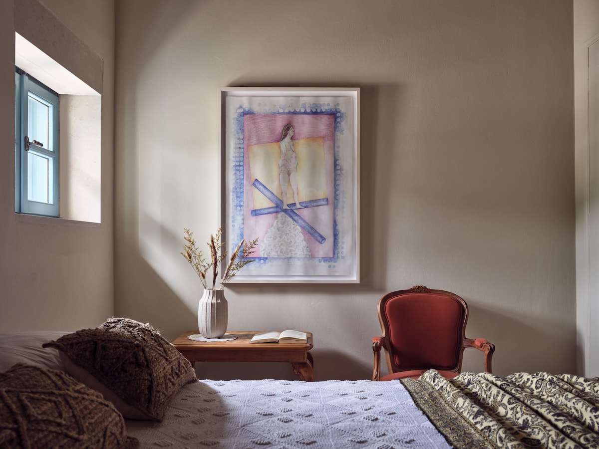 A cozy sleeping area is presented with a spacious bed dressed in textured bedding. A vintage red armchair is positioned beside a small wooden table, which holds an open book and a vase displaying dried flowers. Artwork adorns the wall, and a window allows natural light to enter.