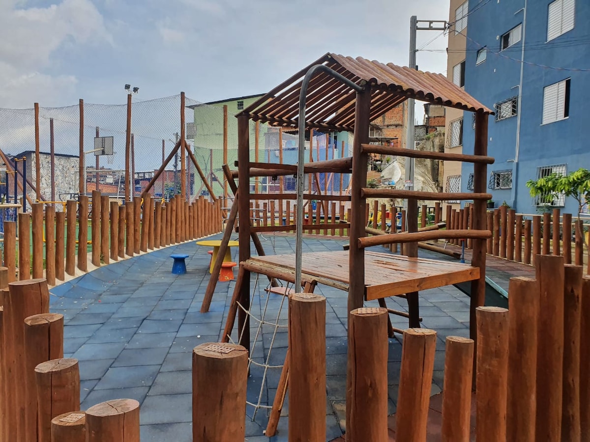 An outdoor play area is featured, surrounded by wooden fencing and logs. A wooden play structure with a roof is visible, alongside colorful benches and pathways. The ground is covered in safety mats, making it accessible and suitable for children.