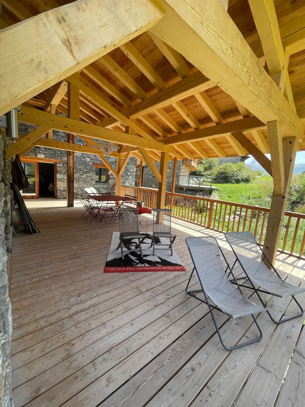 A spacious terrace is shown, featuring a wooden roof supported by beams. Several chairs and tables are arranged, with a textile rug adding comfort to the wooden floor. Lush greenery and distant mountain views are visible beyond the railing, providing a serene outdoor environment.