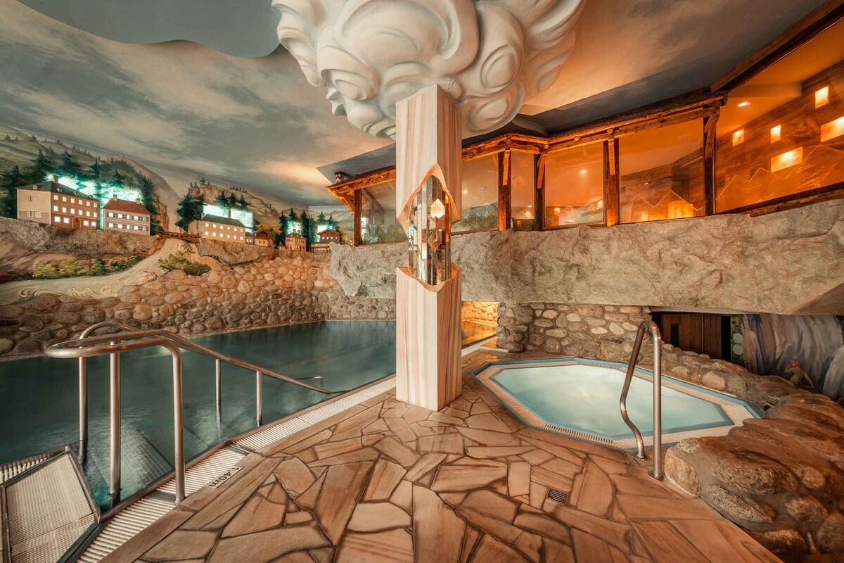 An indoor spa area is showcased with a natural stone design and a tranquil pool. Elegant lighting creates a calming ambiance, while a small hot tub is visible nearby. Cloud-like decor adorns the ceiling, enhancing the serene atmosphere.
