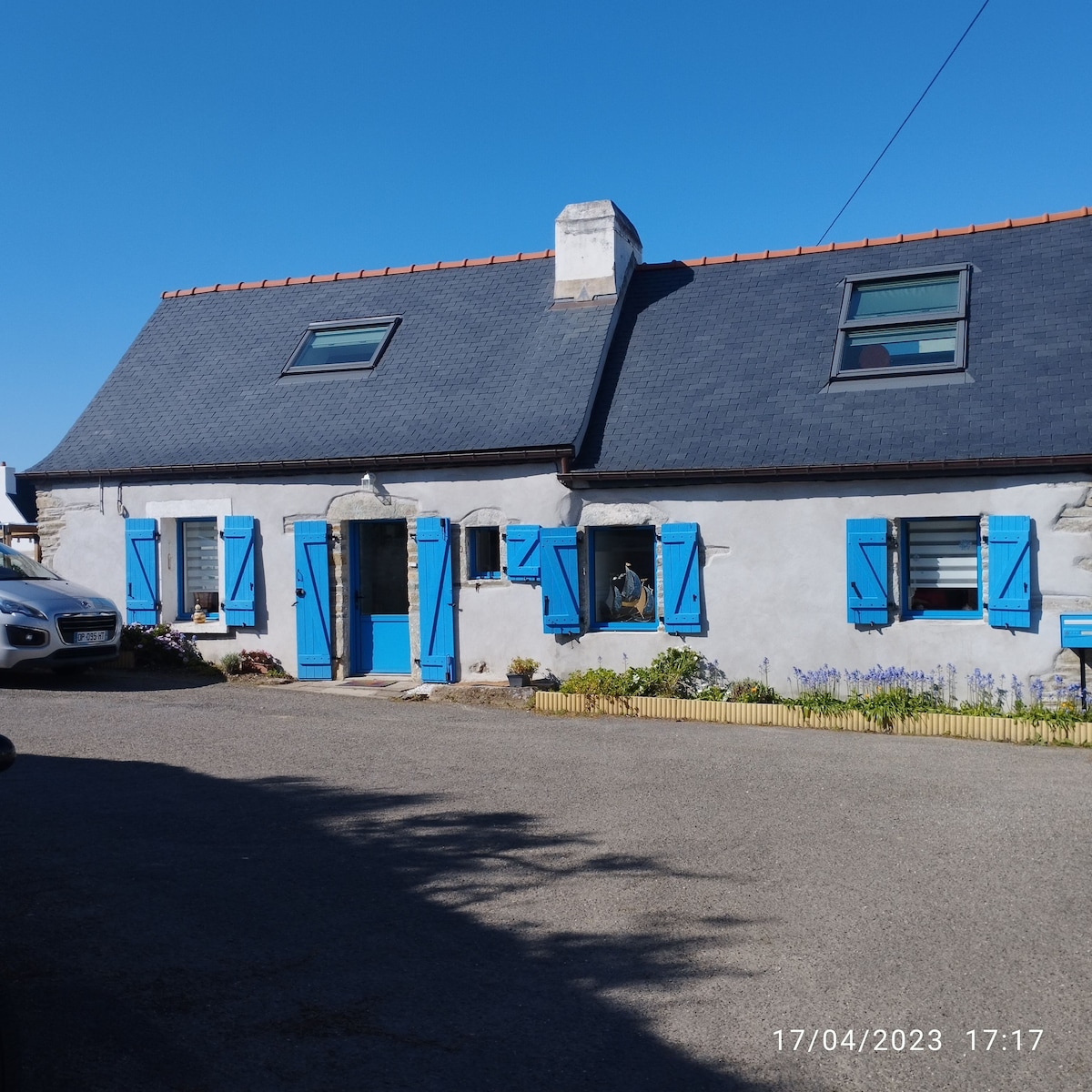 A charming house exterior features a light-colored facade complemented by bright blue window shutters. The roof is dark with visible skylights, and flowers bloom near the base of the building, adding a touch of nature to the entrance. The surrounding area is paved.