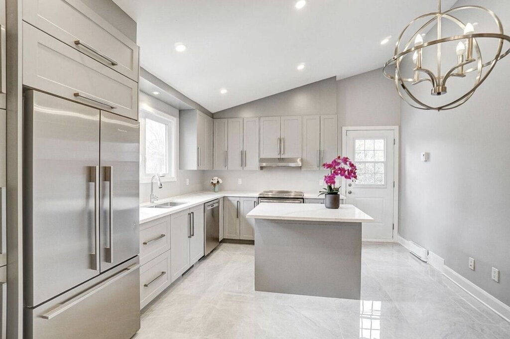 A modern kitchen features sleek cabinetry and stainless steel appliances. A large island is centered, adorned with a potted orchid. Bright lighting accentuates the marble flooring, while a window allows natural light to illuminate the space. A door leads to the exterior.
