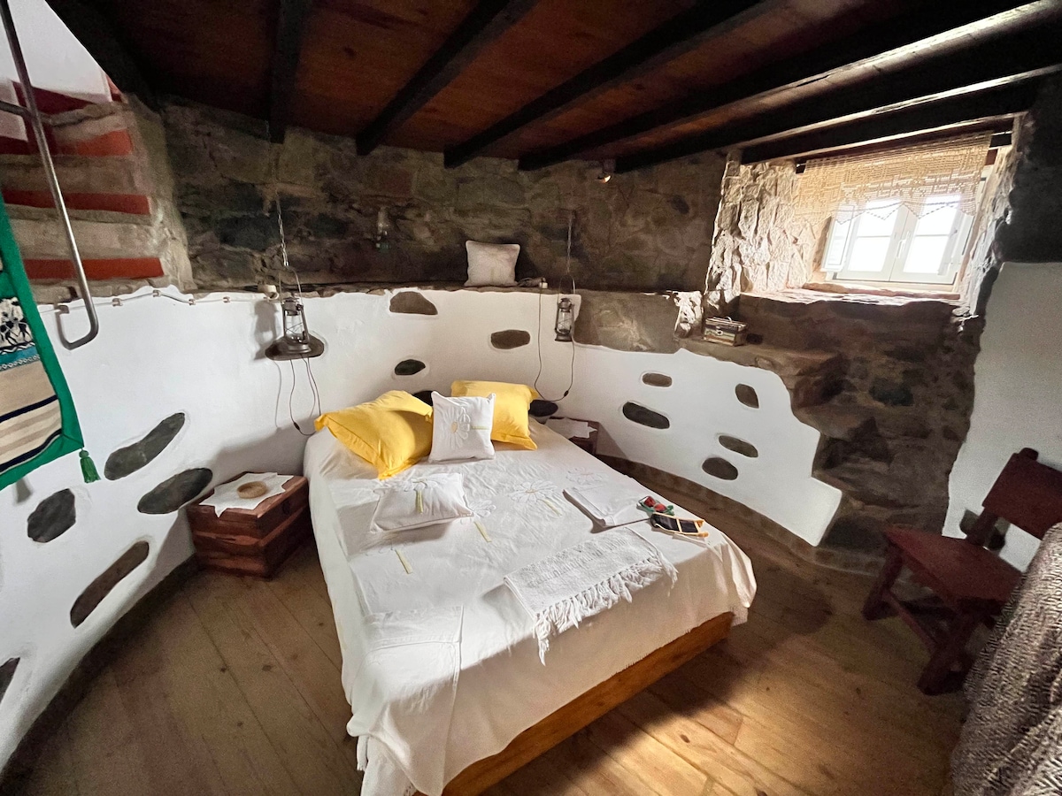 A cozy sleeping area features a spacious bed adorned with a white blanket and yellow pillows. Exposed stone walls create a rustic atmosphere, while natural light enters from a small window. A wooden table and chair complement the simple decor.