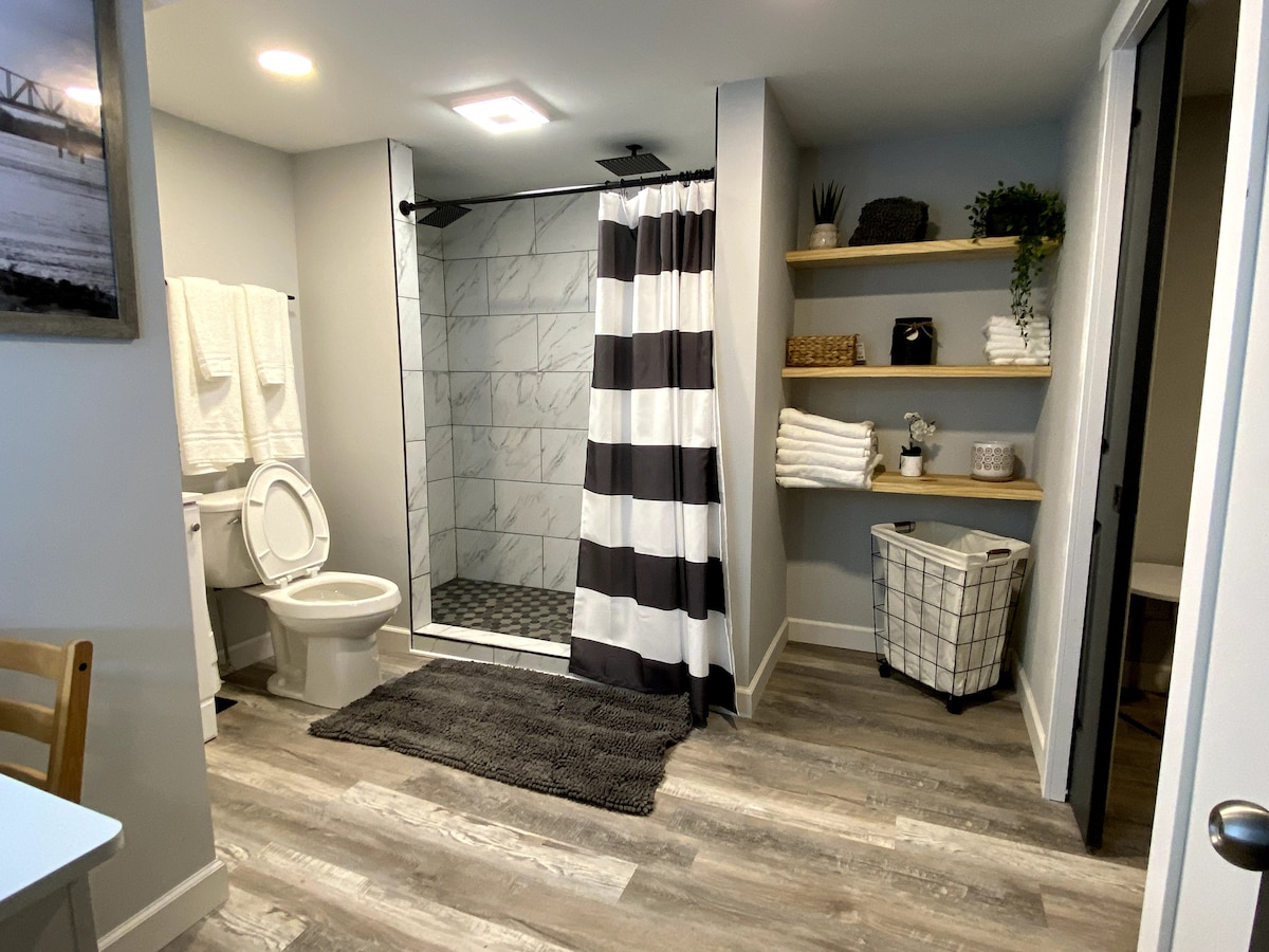 A well-appointed bathroom features a glass shower with a black and white striped curtain. Towel racks hold neatly stacked white towels. A laundry basket and additional storage shelves provide ample space for toiletries and personal items, while soft lighting adds to the overall ambiance.