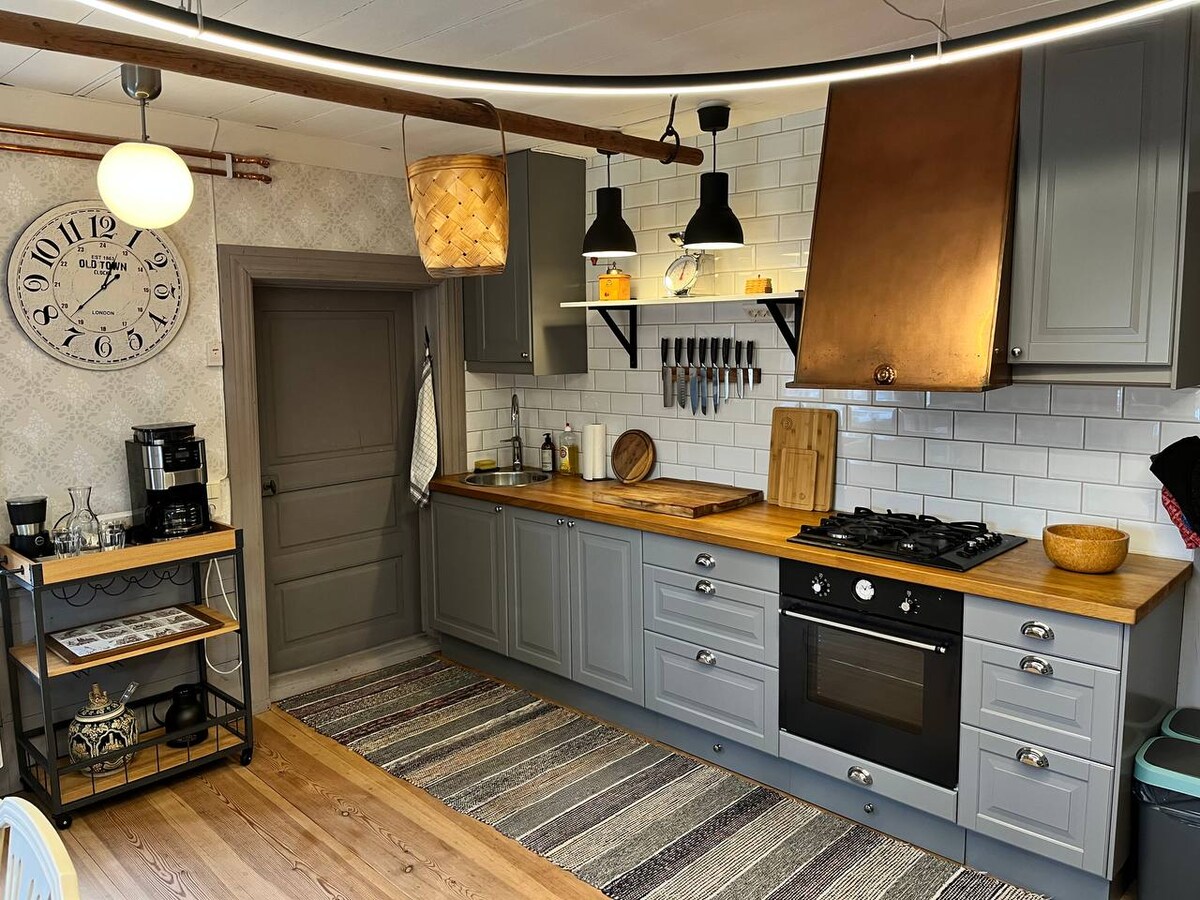 The kitchen features grey cabinetry with a wooden countertop, equipped with essential appliances including a coffee maker and a gas stove. A large wall clock and decorative utensils add character. A kitchen island on wheels provides additional workspace and storage, with a soft rug underfoot.
