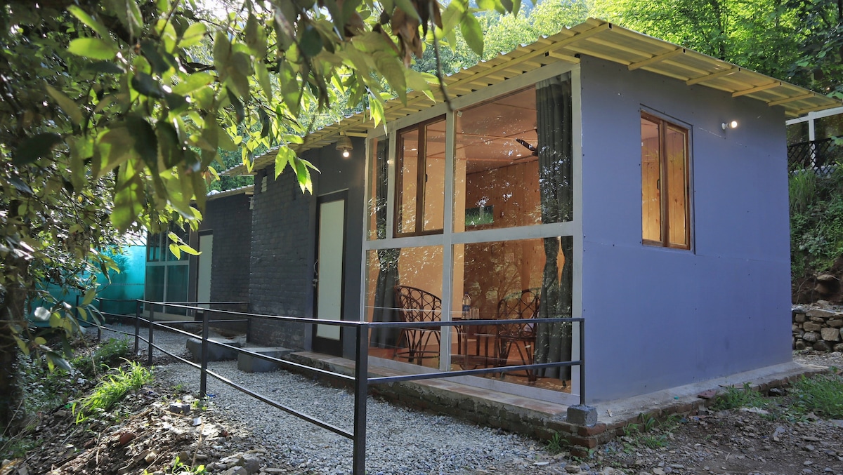 A contemporary cottage is nestled among greenery, featuring large windows that invite natural light. A gravel pathway leads to the entrance, with a small ramp for accessibility. The inviting outdoor space surrounds the structure, enhancing the sense of connection with nature.