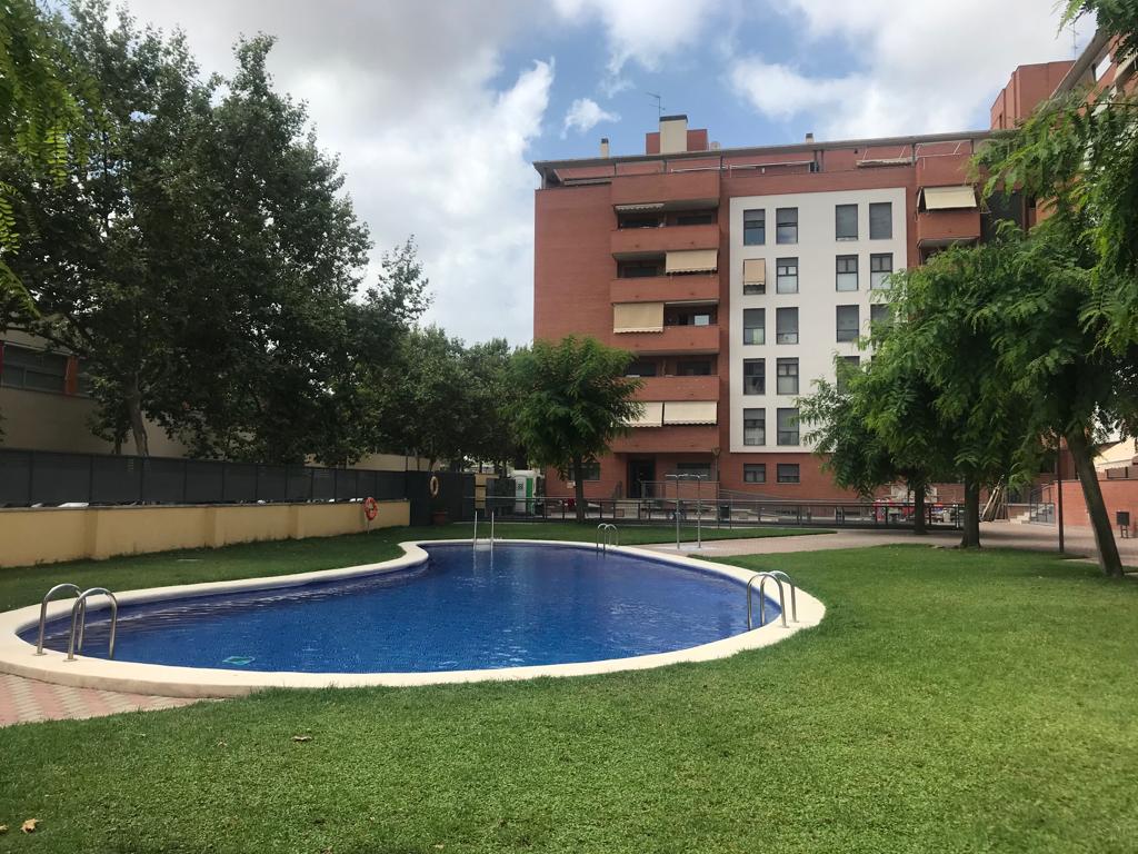 A refreshing swimming pool is set within a green lawn area, surrounded by trees. The pool features a gently curved shape and steps leading into the water. In the background, a multi-story building is visible, complemented by additional greenery.