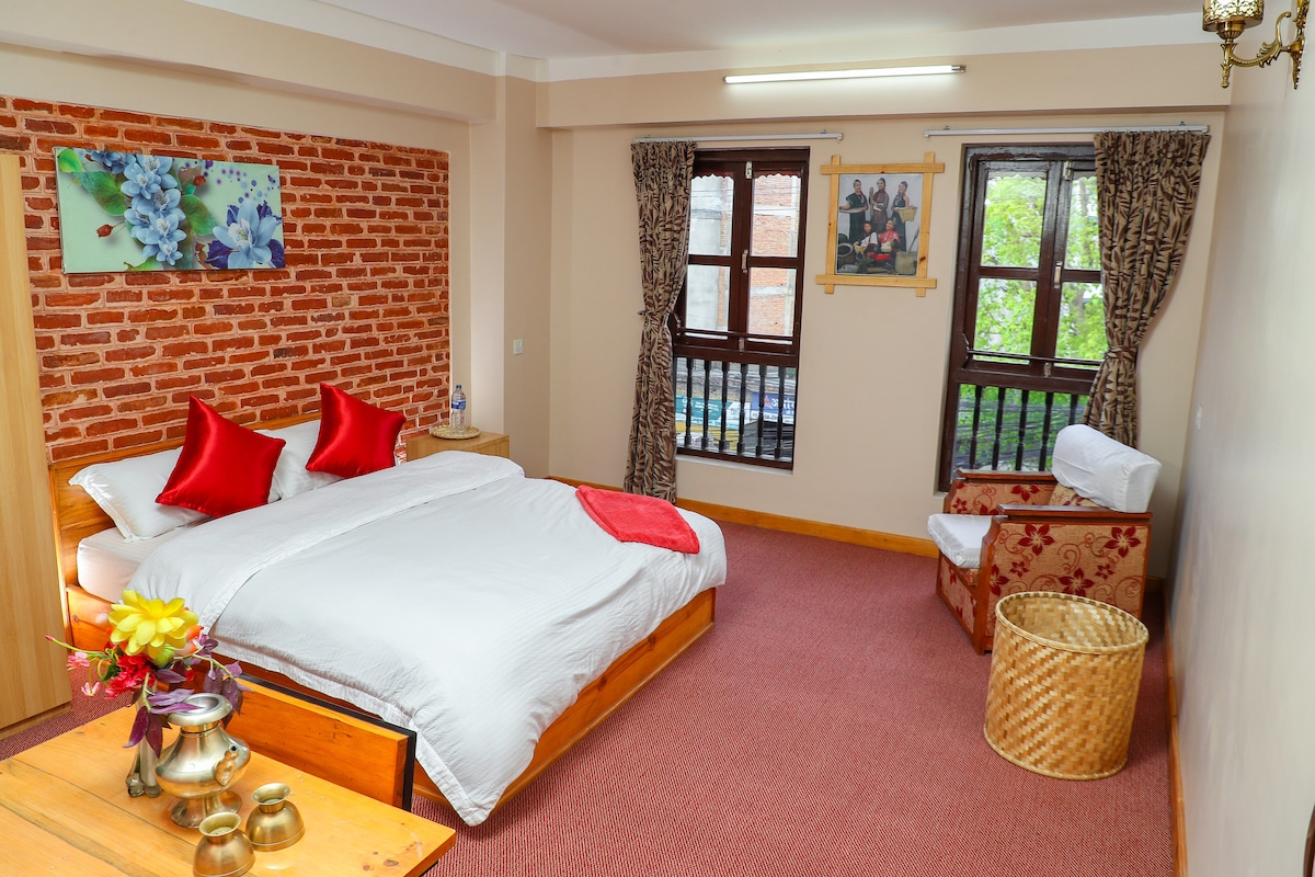 The studio features a clean, uncluttered space with a spacious bed adorned with bright red decorative cushions. Natural light streams through two windows, framed by patterned curtains, illuminating the exposed brick wall and tasteful artwork. A cozy chair and a decorative basket add warmth to the room.