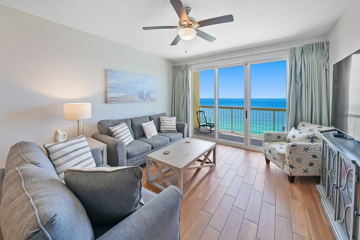 A spacious living area features comfortable grey sofas and a wooden coffee table, complemented by a decorative armchair. Large sliding glass doors provide expansive views of the Gulf, with bright blue water visible beyond the balcony. Light-colored drapes frame the windows.
