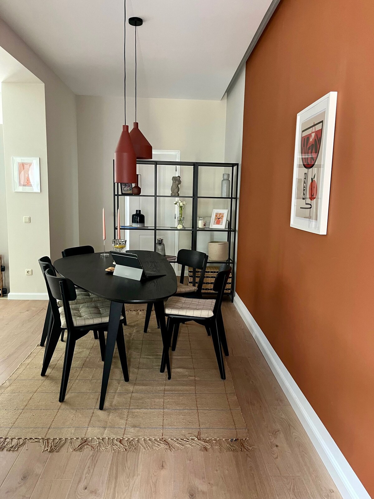 A modern dining area features an oval black table surrounded by four black chairs with light-colored seats. A glass display cabinet filled with decorative items is positioned along the wall. The wall is painted a warm terracotta shade, complemented by framed artwork.