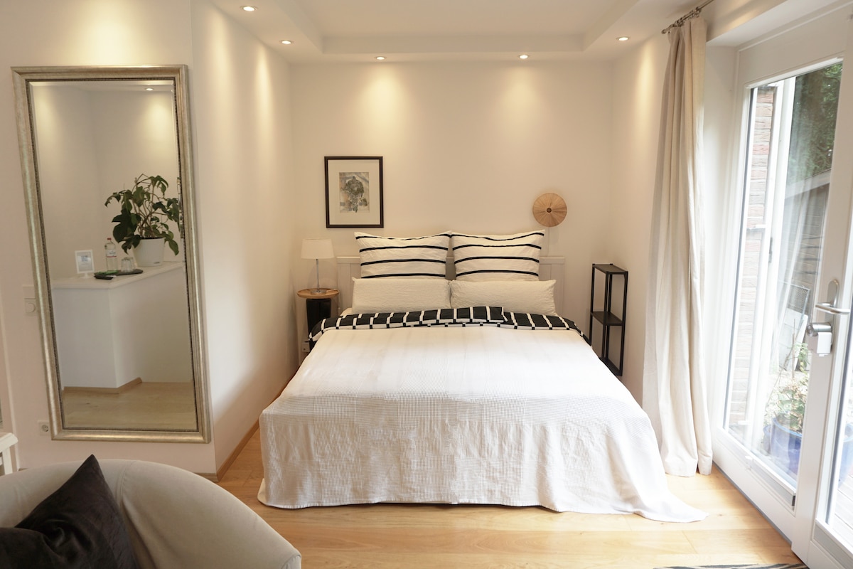 A bright and airy sleeping area features a spacious bed adorned with striped linens. Natural light enters through large glass doors, illuminating the wooden floor. A mirror on the side reflects the minimalist design, with potted plants and bedside lamps enhancing the decor.