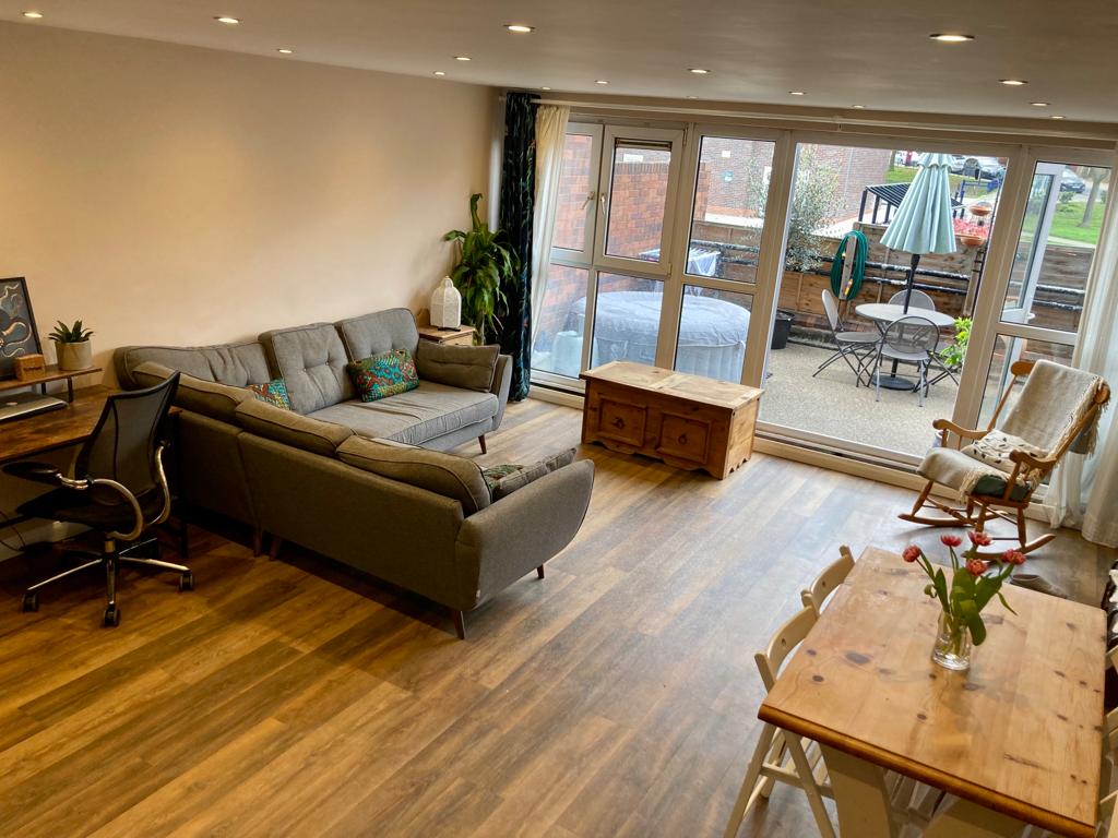 The open-plan living area features a comfortable gray sofa and a wooden dining table with chairs. Large glass doors provide natural light and lead to the courtyard garden. A potted plant adds a touch of greenery to the space, enhancing the inviting atmosphere.