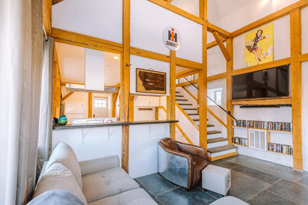 An inviting living area is characterized by exposed wooden beams and a modern aesthetic. A spacious couch is positioned adjacent to a sleek coffee table. A staircase leads to the upper level, while a flat-screen television is mounted on the wall, accompanied by a collection of DVDs on a nearby shelf.