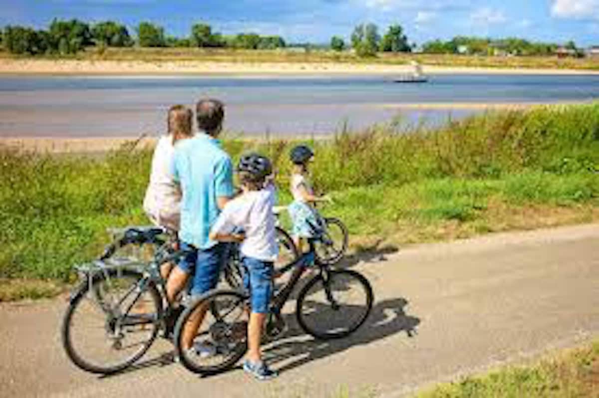 A family is seen on bicycles along a path by the river, with greenery surrounding the area. The scenic view reveals flowing water and trees in the distance under a partly cloudy sky. Safety helmets are worn by the children.