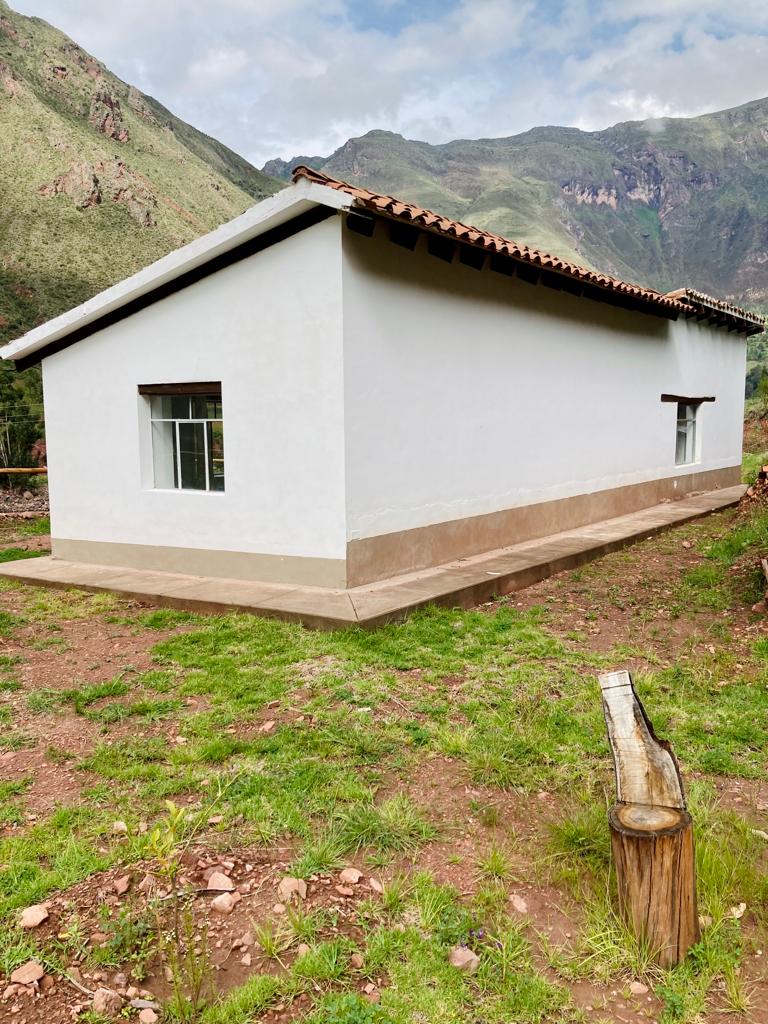 A simple white structure is situated on a grassy area, surrounded by lush green hills. The building features a prominent window on the side, providing natural light. A rustic wooden water feature is visible in the foreground, enhancing the natural setting.