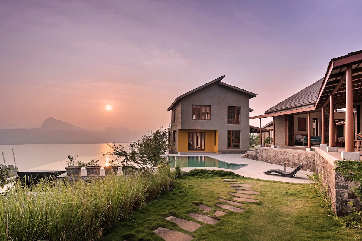 A modern villa is highlighted against a sunset over Pawna Lake. A sleek infinity pool is visible, complemented by a stone pathway leading through lush greenery. Expansive windows showcase the scenic landscape and the architecture features a blend of earthy tones.