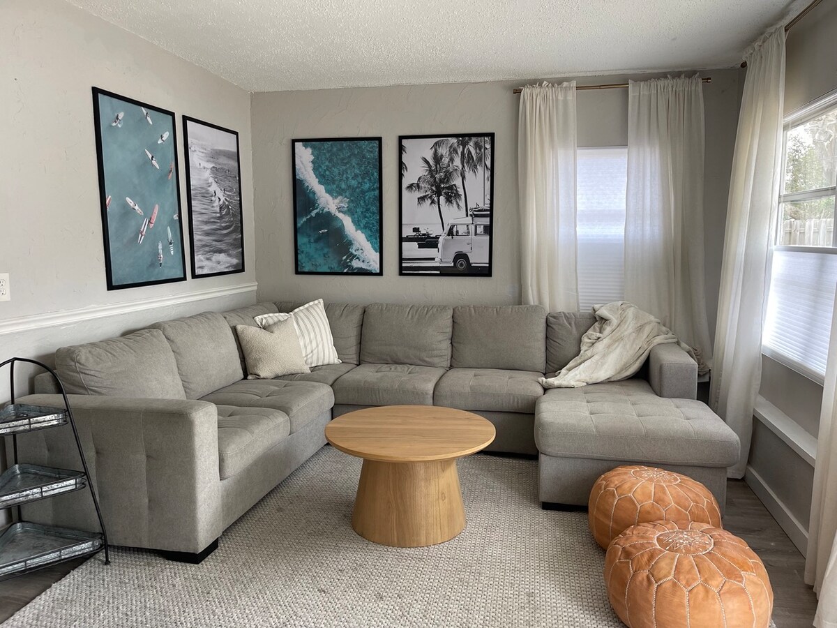 A spacious living room features a large sectional sofa in neutral tones, complemented by a round wooden coffee table. Four framed artworks depicting coastal scenes adorn the walls. Light enters through white curtains, and a textured area rug covers the floor, enhancing the cozy environment.