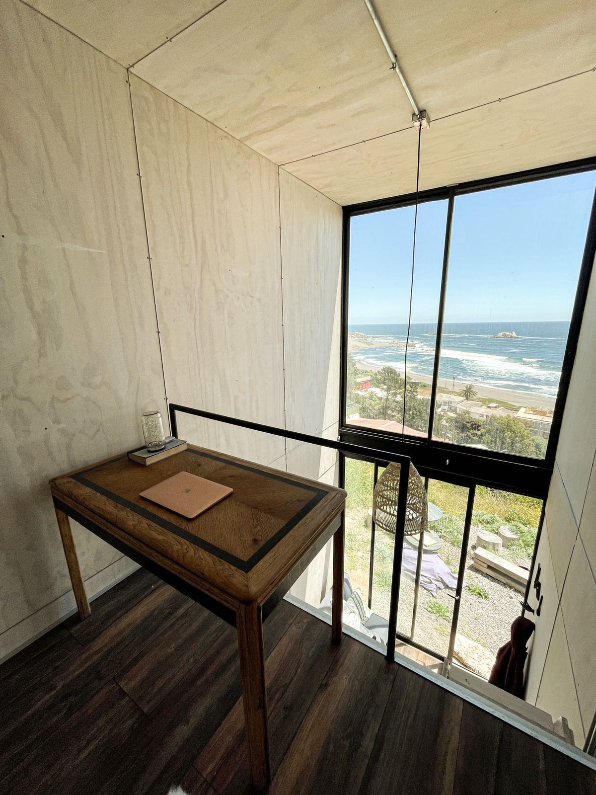 A bright work corner is positioned near a large window, showcasing panoramic views of the ocean and coastline. A wooden table with a glass of water and a notebook is visible, inviting relaxation and focus while enjoying the scenery.