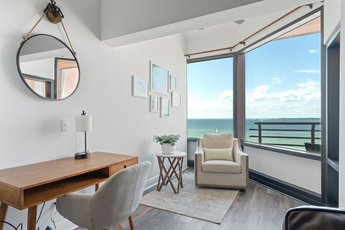 A corner of the living area is presented with large windows offering expansive views of the water. A wooden desk and a light-colored chair provide a workspace, while a soft armchair and small table create a cozy seating arrangement. Natural light floods the space.