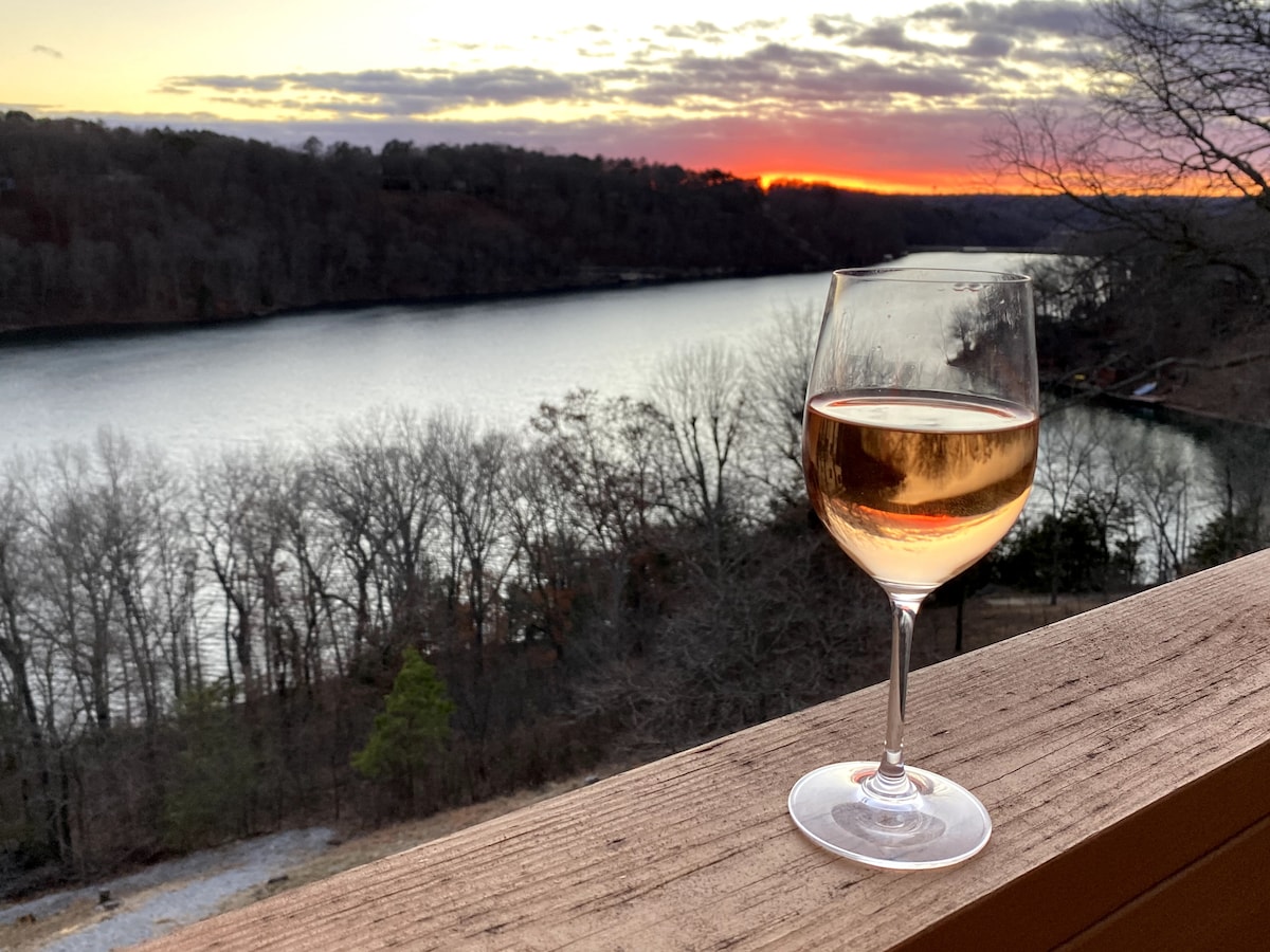 A glass of rosé sits on a wooden railing, overlooking a serene lake at sunset. Soft hues of orange and purple are reflected in the water, framed by leafless trees lining the shore, creating a tranquil atmosphere.