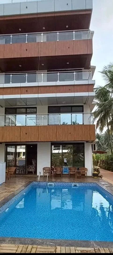 A modern building with wooden accents features multiple balconies overlooking a clear blue pool. The outdoor area includes seating arrangements near the pool, surrounded by lush greenery. Large glass windows allow for views of the interior spaces, enhancing the connection to the outdoor environment.