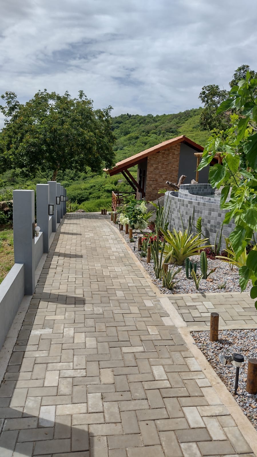 A well-maintained pathway made of gray pavers leads toward a rustic building, flanked by green vegetation and various plants. The scene is set against a backdrop of hills, creating a natural and serene atmosphere.