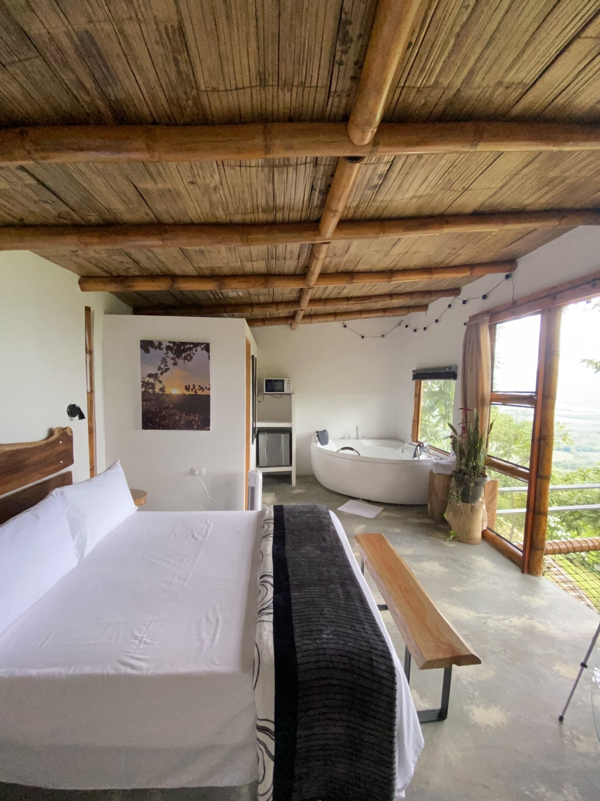A spacious cabin interior is showcased, featuring a comfortable bed with neatly arranged linens. A freestanding bathtub is positioned near large windows, allowing natural light to illuminate the space. A wooden bench adds a rustic touch, complemented by bamboo-style ceiling beams.