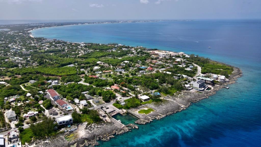 An aerial view captures the coastal landscape, displaying lush greenery alongside residential areas and a mix of single-standing homes and condos. The clear blue ocean meets rocky shorelines, with a glimpse of the beach in the distance, showcasing the vibrant colors of the Cayman Islands.