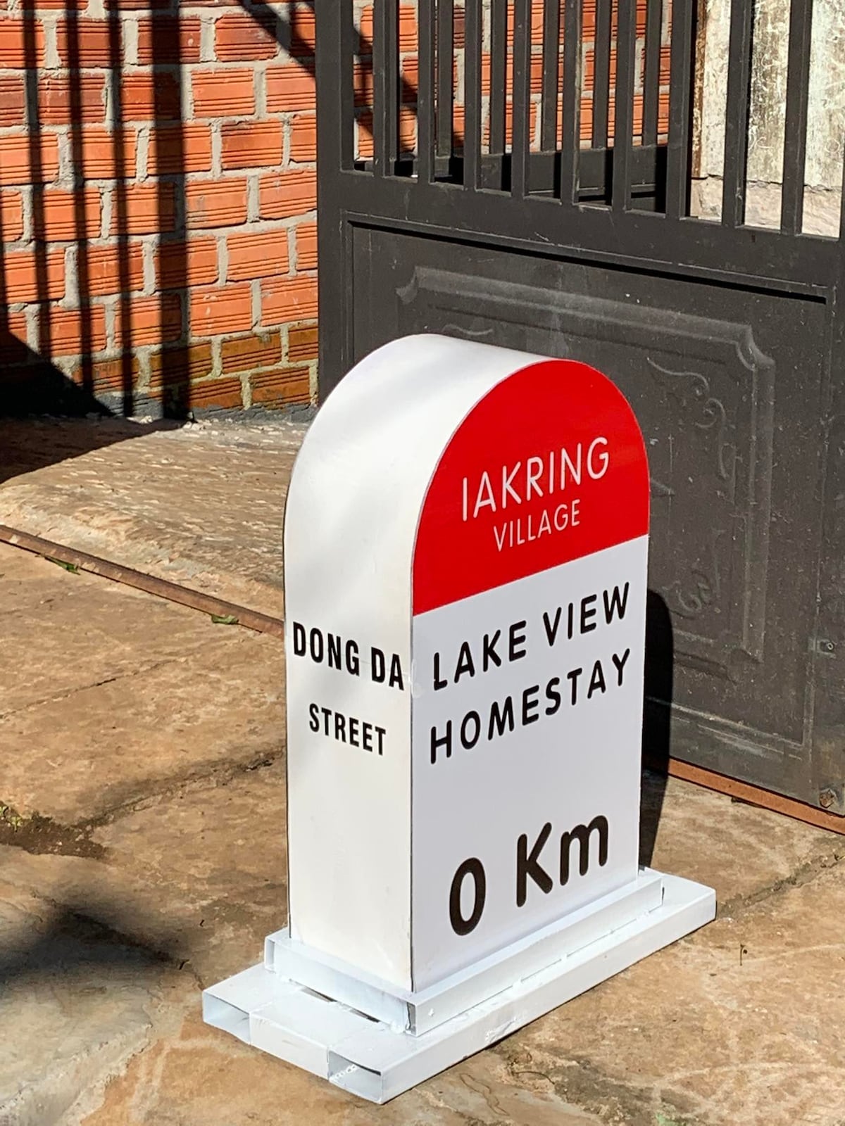 A rectangular white sign with a rounded top is located at the entrance. The sign displays 'LAKE VIEW HOMESTAY' and '0 Km' prominently, with 'DONG DA STREET' beneath. A brick wall and stone pathway are visible in the background.