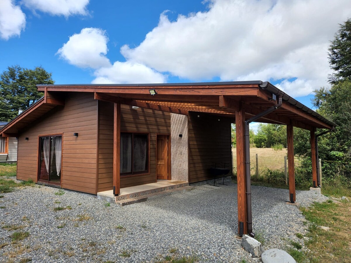 A single-story cabin is displayed, featuring a wooden exterior and a covered front porch. Large windows are present, allowing natural light to brighten the interior. A gravel pathway leads up to the entrance, surrounded by a grassy area and trees in the background.