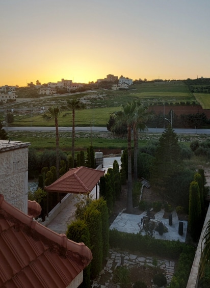 An expansive garden is visible, framed by lush greenery and lined with palm trees. The sun sets in the background, casting a warm glow over the landscape, which includes gently rolling hills and distant buildings. A path leads through the garden, enhancing the serene environment.