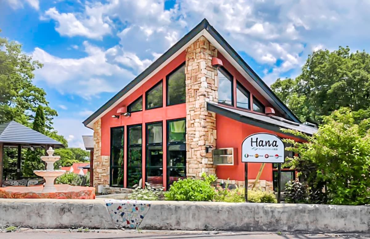 A spacious building featuring large glass windows allows natural light to fill the interior. The exterior is a blend of stone and wood, with a welcoming sign displaying 'Hana'. Surrounding greenery and a decorative fountain add to the inviting atmosphere.