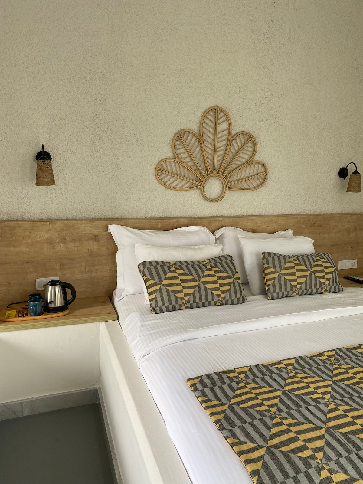 A well-appointed sleeping area is highlighted by a spacious bed with patterned pillows and a textured throw. Wood accents are present on the headboard and bedside shelf, which holds a kettle and cups, contributing to a cozy atmosphere.