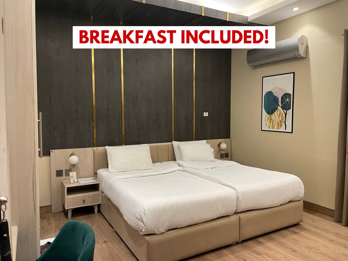A spacious guest room features two separate beds with white linens, accented by a neutral-toned headboard. Wall-mounted art adds a pop of color, while ambient lighting creates a comfortable setting. Air conditioning is visible on the wall, and modern furnishings complete the inviting ambiance.
