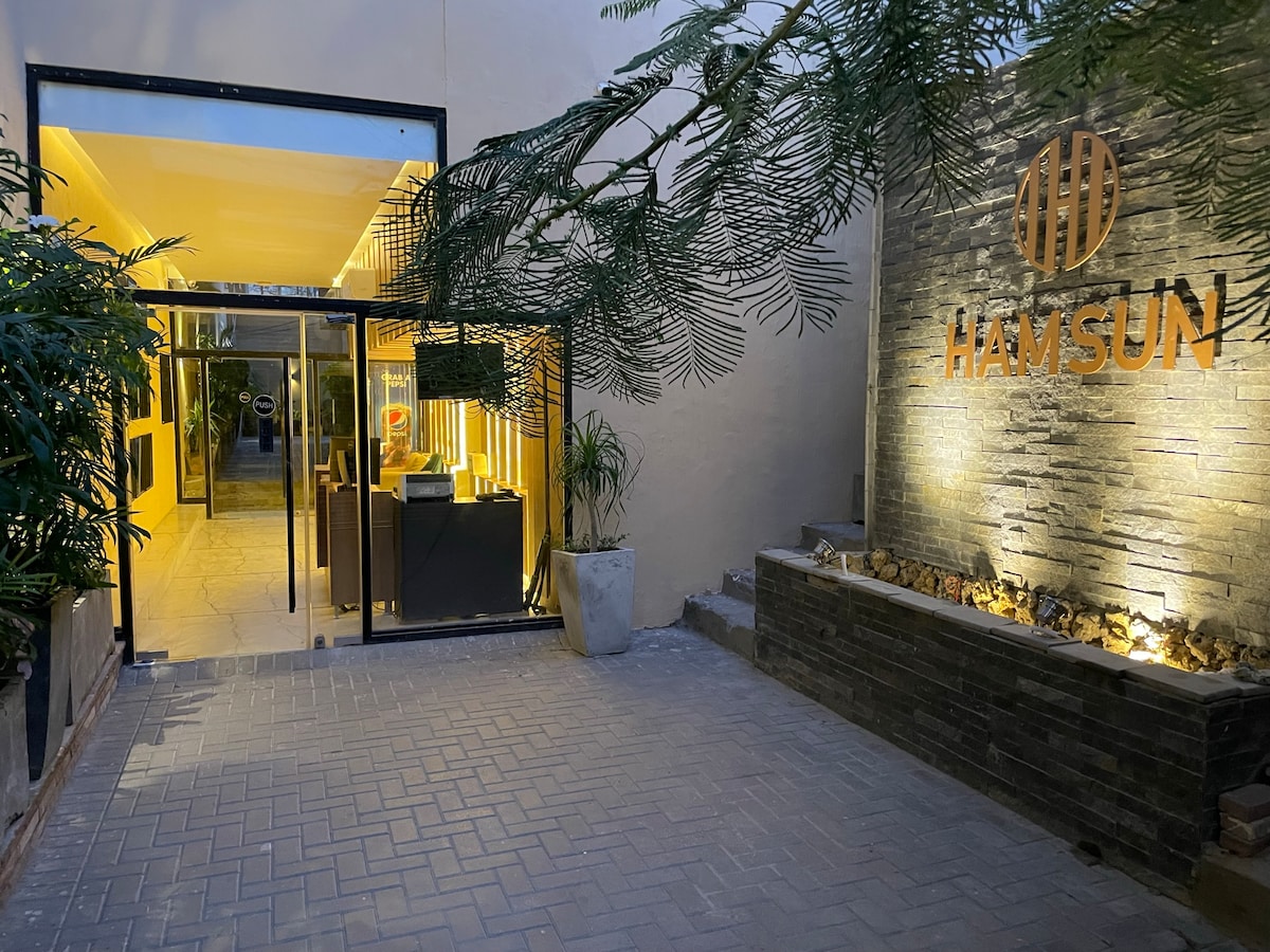 The entrance to the property is illuminated, featuring a sleek glass door that opens to a well-maintained pathway. Lush greenery frames the entrance and a stylish sign is displayed prominently on the wall. Soft lighting enhances the welcoming ambiance.