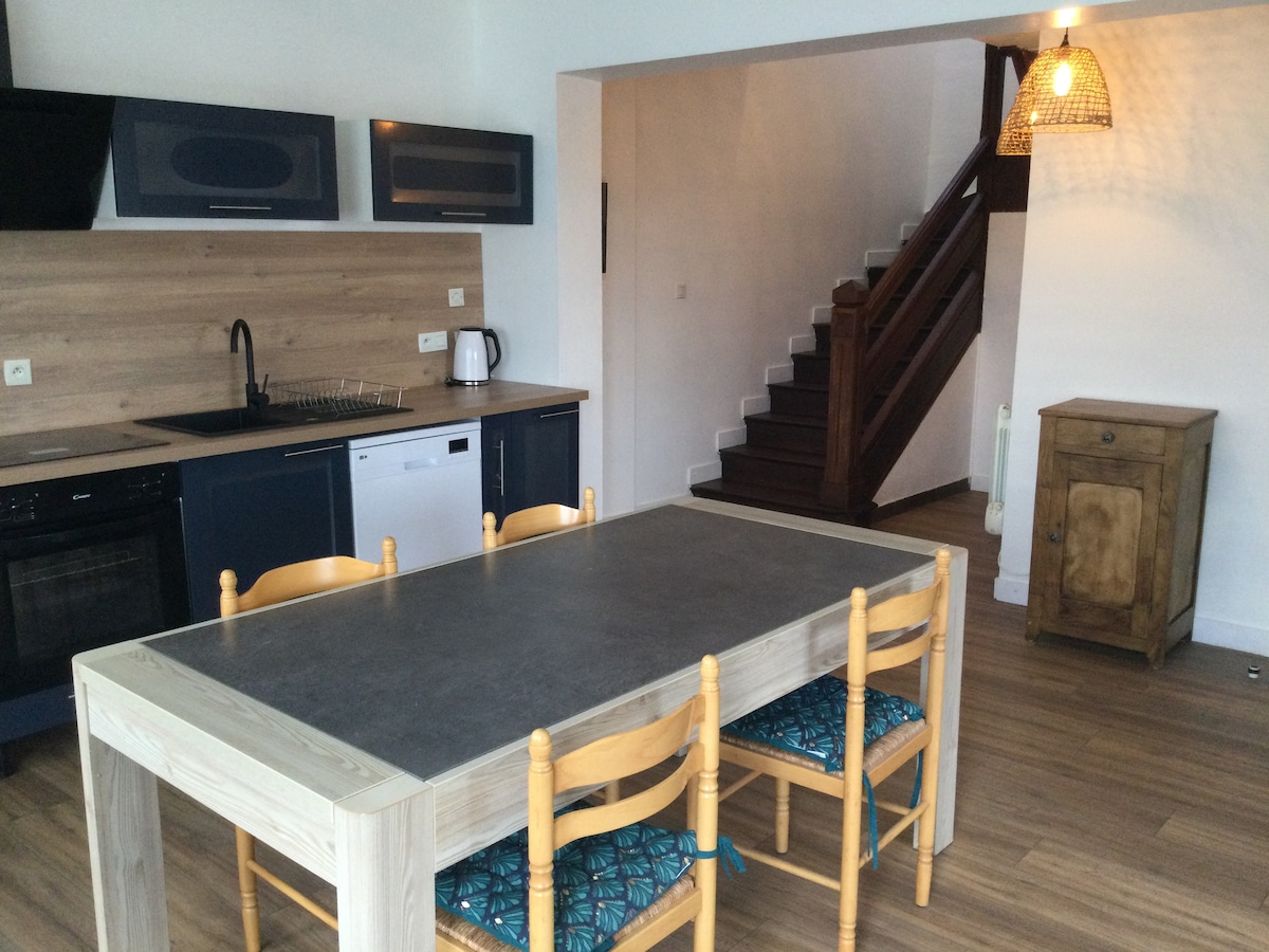 An open kitchen area is visible, featuring a modern design with dark cabinetry and a wood accent wall. A spacious dining table with wooden chairs is positioned centrally. A staircase leading to the upper floor can be seen in the background, enhancing the room's functional layout.
