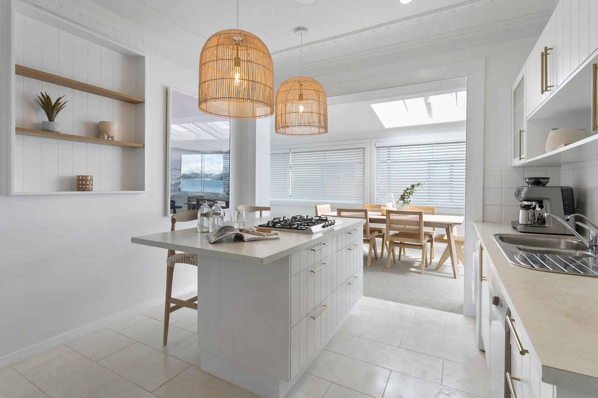 A modern kitchen features a central island with light-colored cabinetry and a gas stove. Two pendant lights hang above, providing soft illumination. Natural light enters through large windows, highlighting a nearby dining area with wooden furniture, creating a bright and inviting setting.