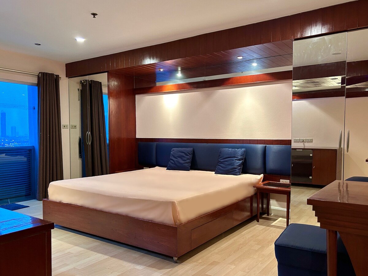 A spacious bedroom features a large bed with a cream-colored coverlet and dark blue pillows. Mirrored panels reflect the room's wooden accents. Soft lighting highlights the elegance of the space, while large windows offer views of the city beyond.