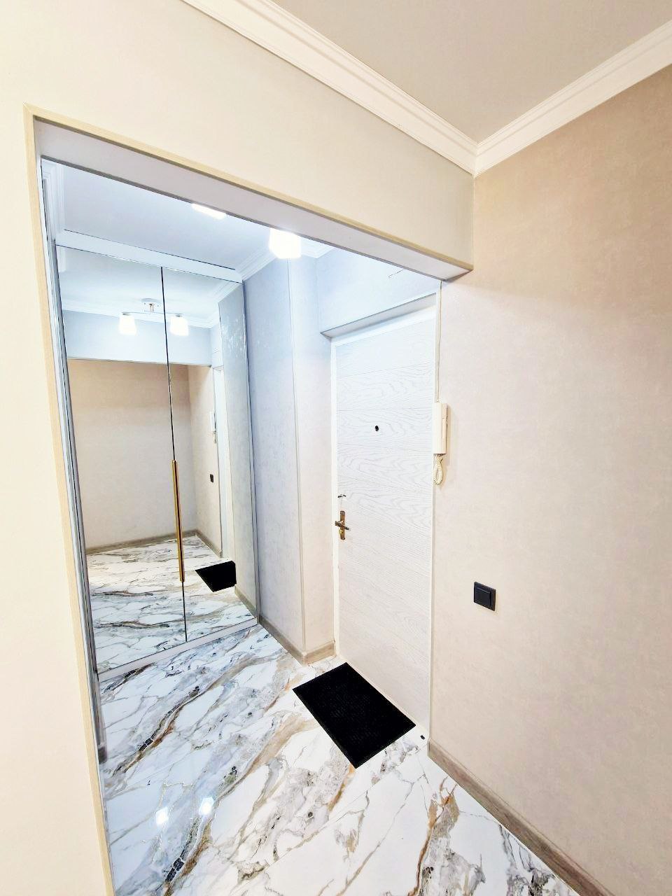 The entryway features sleek, marble flooring that leads to a white door. A full-length mirror is mounted on one wall, reflecting the light and enhancing the space. A small black mat is placed at the entrance.