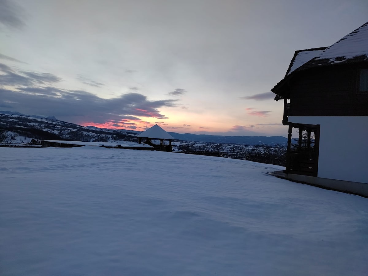 A serene winter landscape is captured with a snow-covered ground extending towards the horizon. A wooden structure is visible on the right, while the sky displays gentle hues of pink and purple from the sunset. Rolling hills fade into the distance, enhancing the tranquil setting.