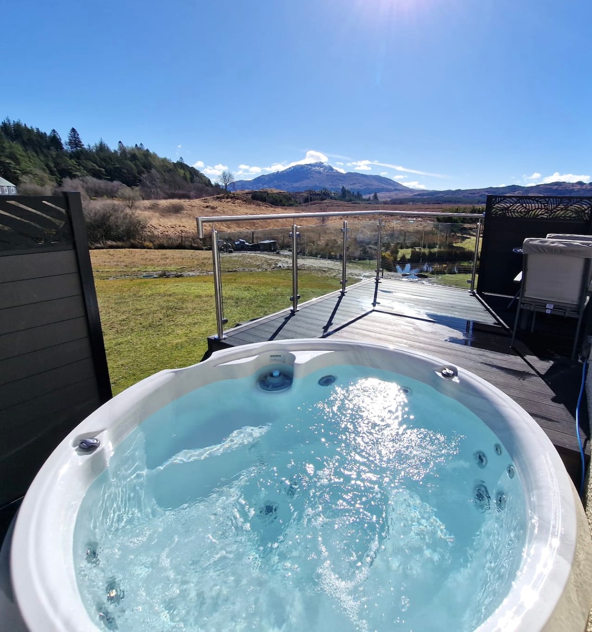 A hot tub is positioned on a deck, offering views of the surrounding landscape and hills. The tranquil water is visible, reflecting the bright blue sky. A glass barrier surrounds the area, enhancing the picturesque outdoor space.